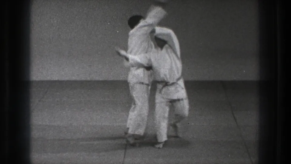 A Seoi Nage throw is underway as one adult in a white gi carries another across his back in the 1971 Tokyo training scene.