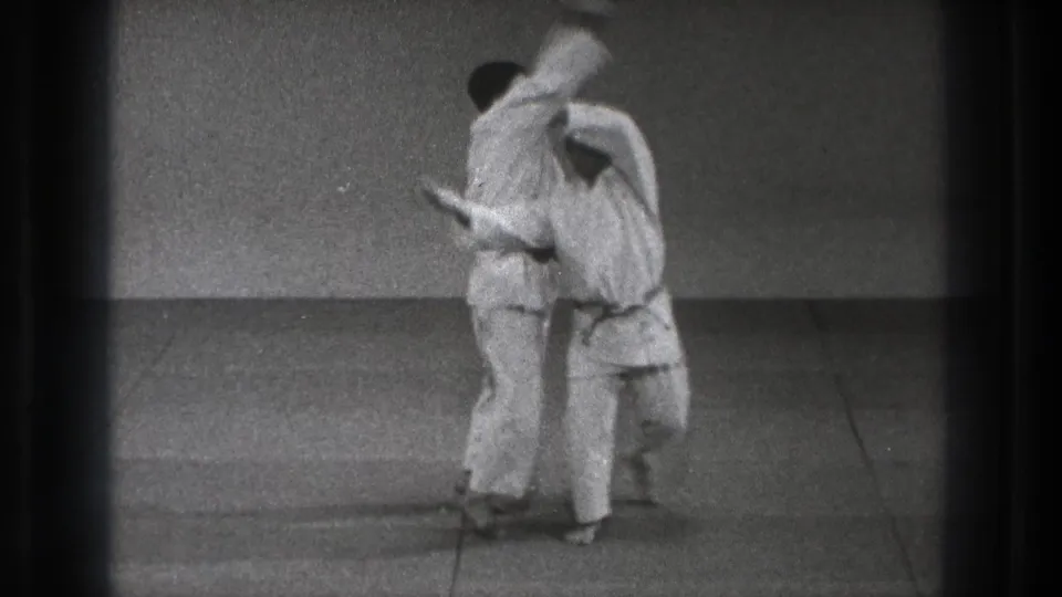 The martial-arts demonstration continues as the thrower supports the other man across his back on a plain training mat.