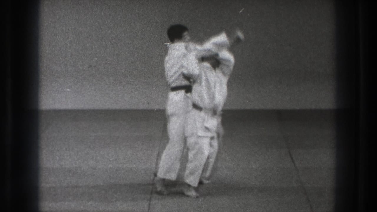 In 1971 Tokyo footage, two adult men in white gi stand close together as a martial arts throw begins on a simple mat.
