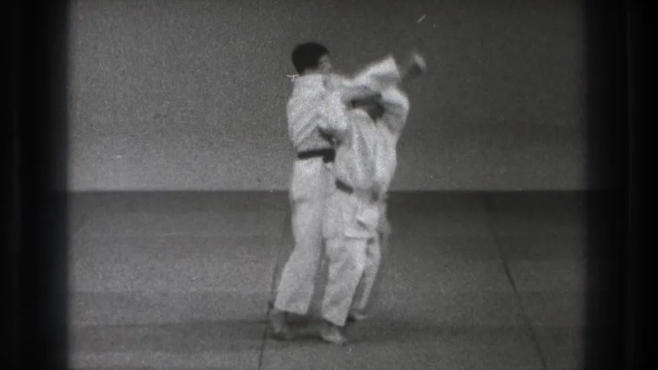 In 1971 Tokyo footage, two adult men in white gi stand close together as a martial arts throw begins on a simple mat.
