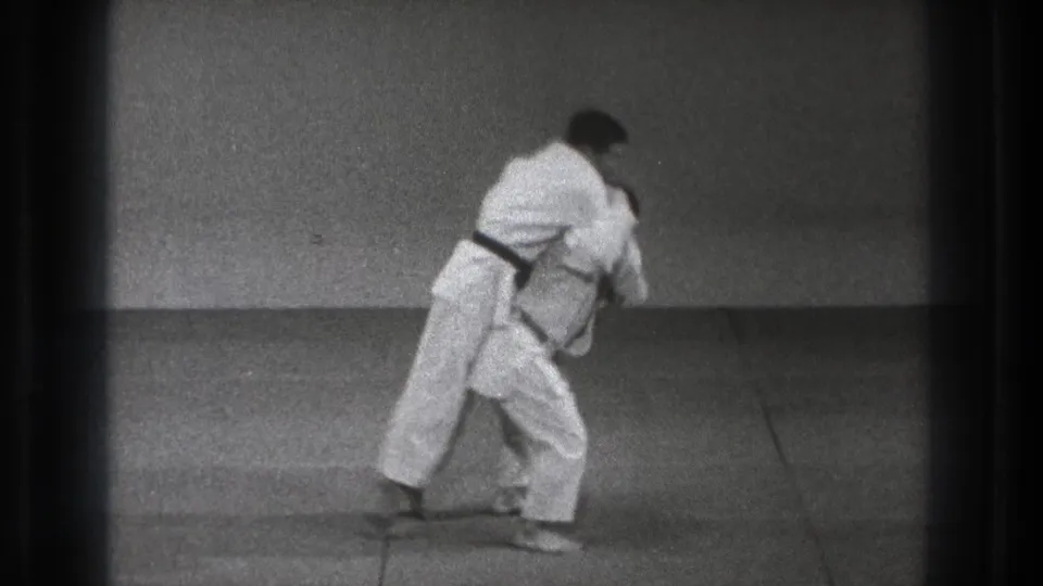 The black-belt practitioner rotates his body into the throw while the second man in a white gi leans forward on the mat.