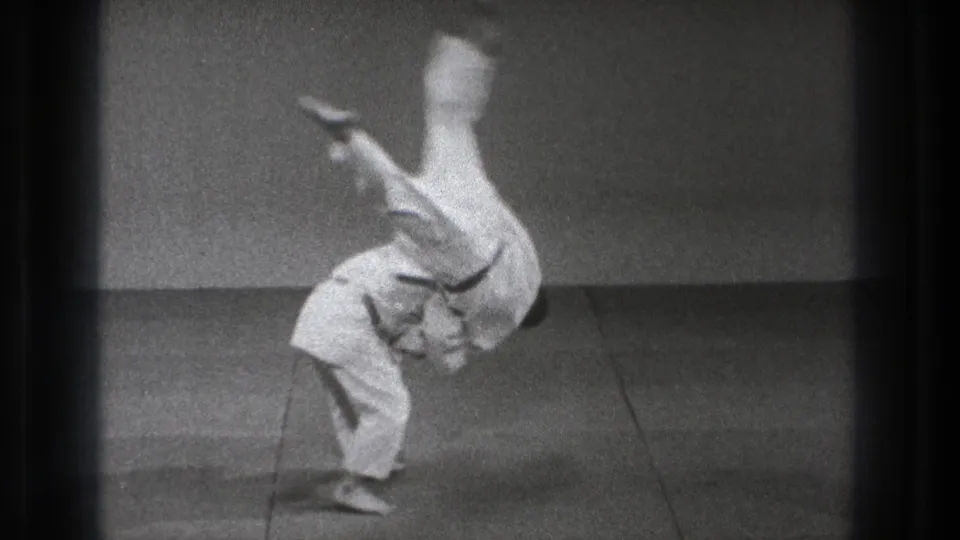 The throw reaches its peak as one man flips upside down over the bent black-belt practitioner on the mat.