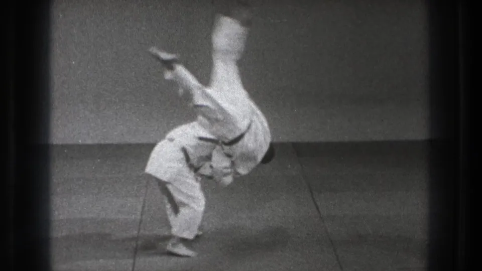 In 1971 Tokyo footage, two karate practitioners in white gi and black belts execute a throw on a plain indoor training mat.