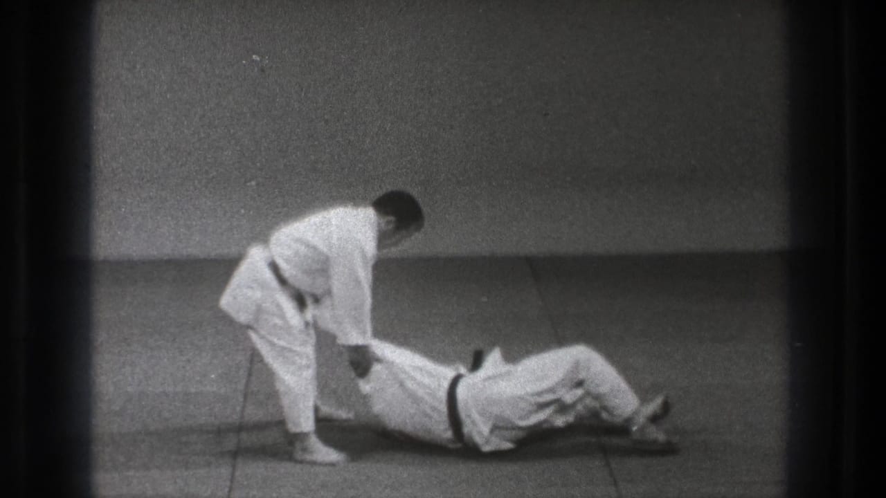 One man lies on the mat while another stands over him holding his arm.