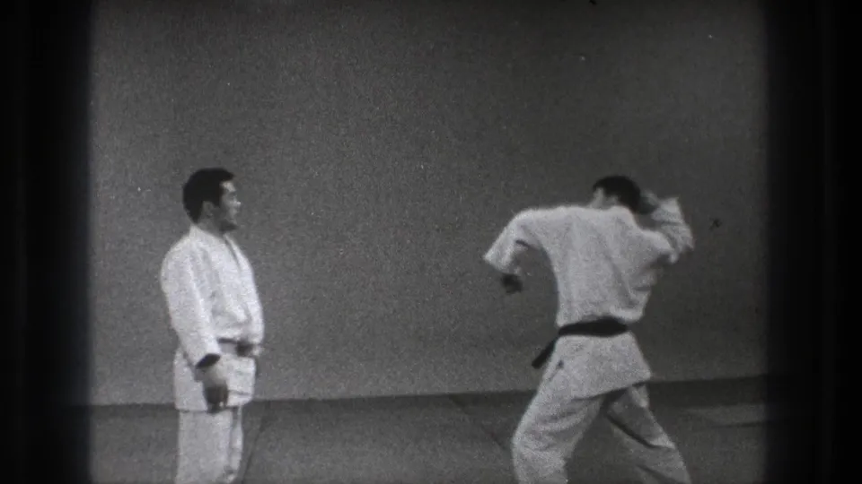 Two karate practitioners face off on a simple indoor mat, with one moving into a strike or block in the 1971 Tokyo footage.