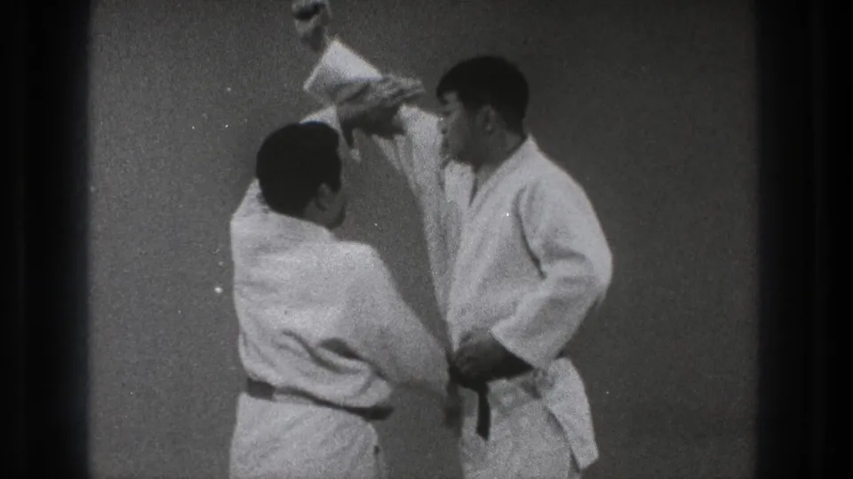 In 1971 Tokyo footage, two adult men in white gi stand close together as one raises an arm to begin a throw.