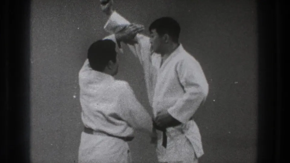 Two adult men in white gi are shown from a home-movie angle as a throwing movement begins in a plain indoor space.