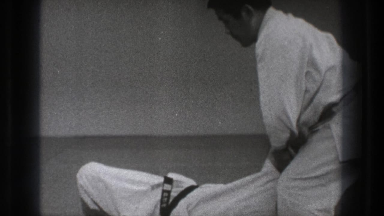 Karate practitioner kneels or reclines while another adjusts position nearby on a mat.