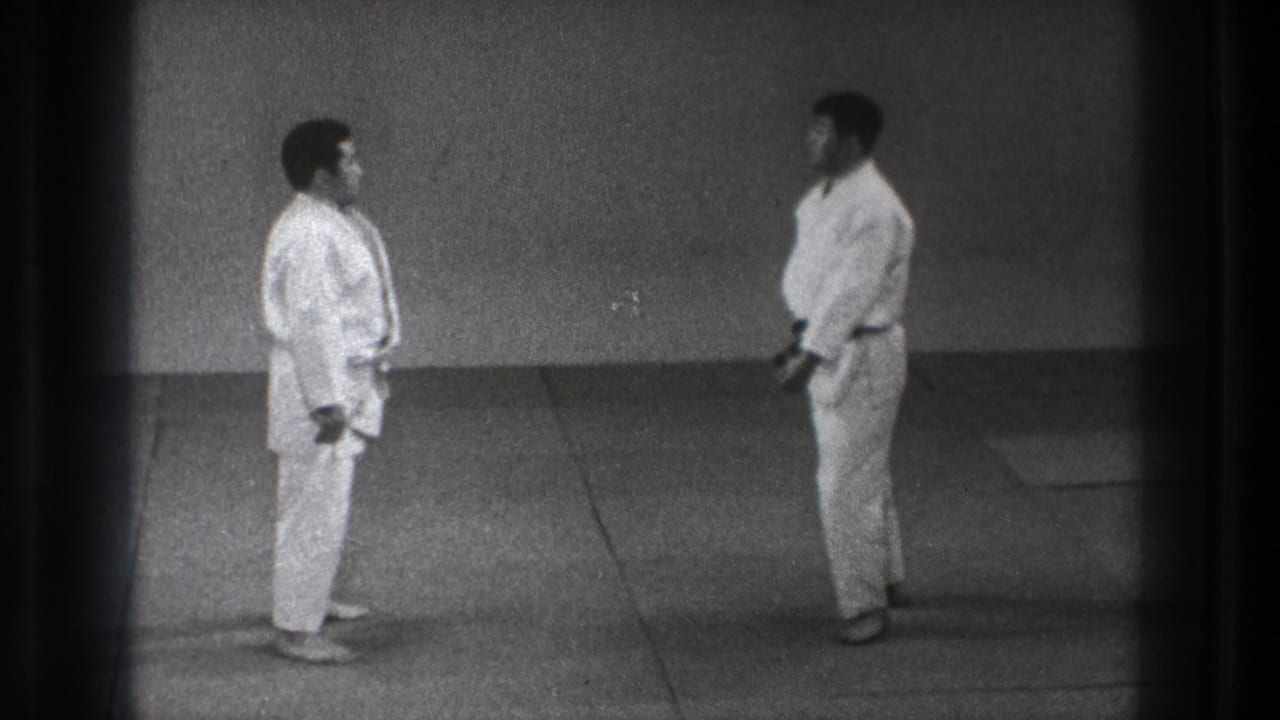 Two karate practitioners stand facing each other on a plain mat surface.