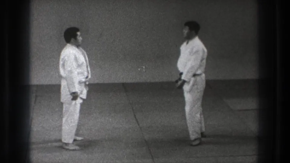 Two karate practitioners in white gi and dark belts face one another in a sparse indoor practice space in the supplied Tokyo 1971 footage.