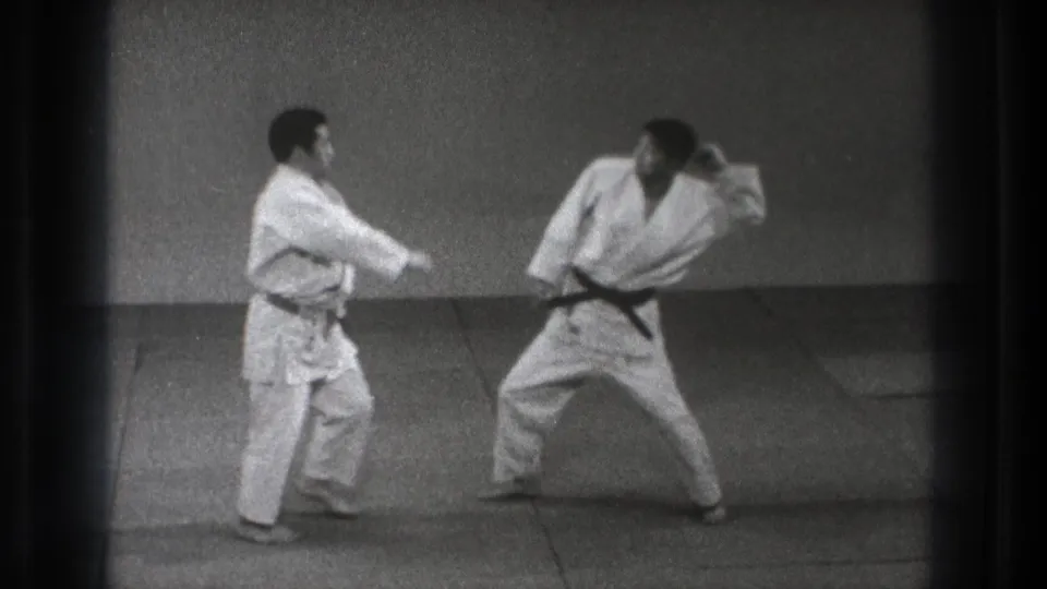In black-and-white 8mm footage, one karate practitioner extends an arm while the other leans away in a controlled sparring motion.