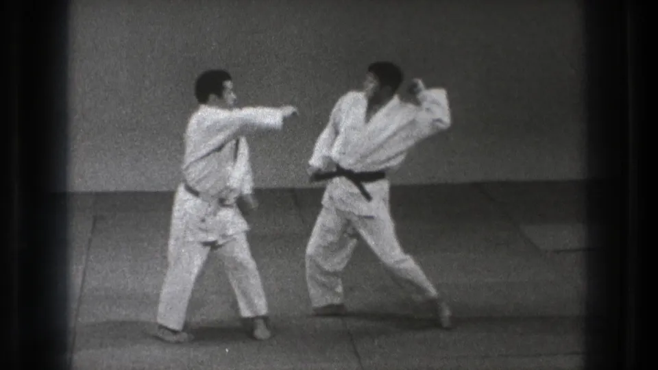 In Tokyo 1971 footage, two karate practitioners in white gi stand in a defensive exchange on a plain indoor mat.