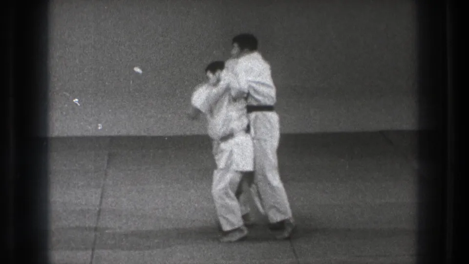 A black-belt karate practitioner appears to control his partner from behind in a Tokyo 1971 indoor training drill.