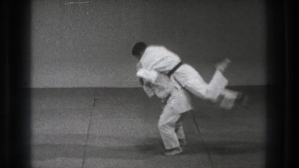 In supported 1971 Tokyo footage, two martial artists in white gi and dark belts start a controlled throw in a dojo-like training space.