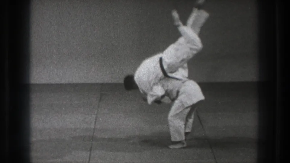 A martial artist is inverted over his partner during a controlled throw in the black-and-white 1971 Tokyo training sequence.