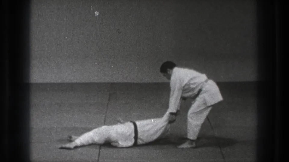 At the end of the throw, one martial artist lies face down on the mat as his partner stands nearby holding a leg.