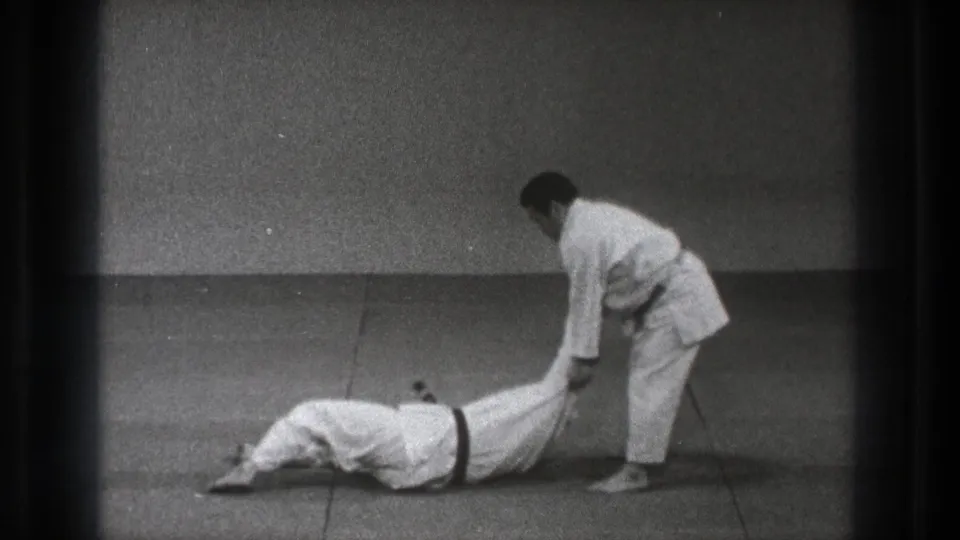 In a 1971 Tokyo dojo scene, one man in a white gi grips another who is down on the tatami after a throw.