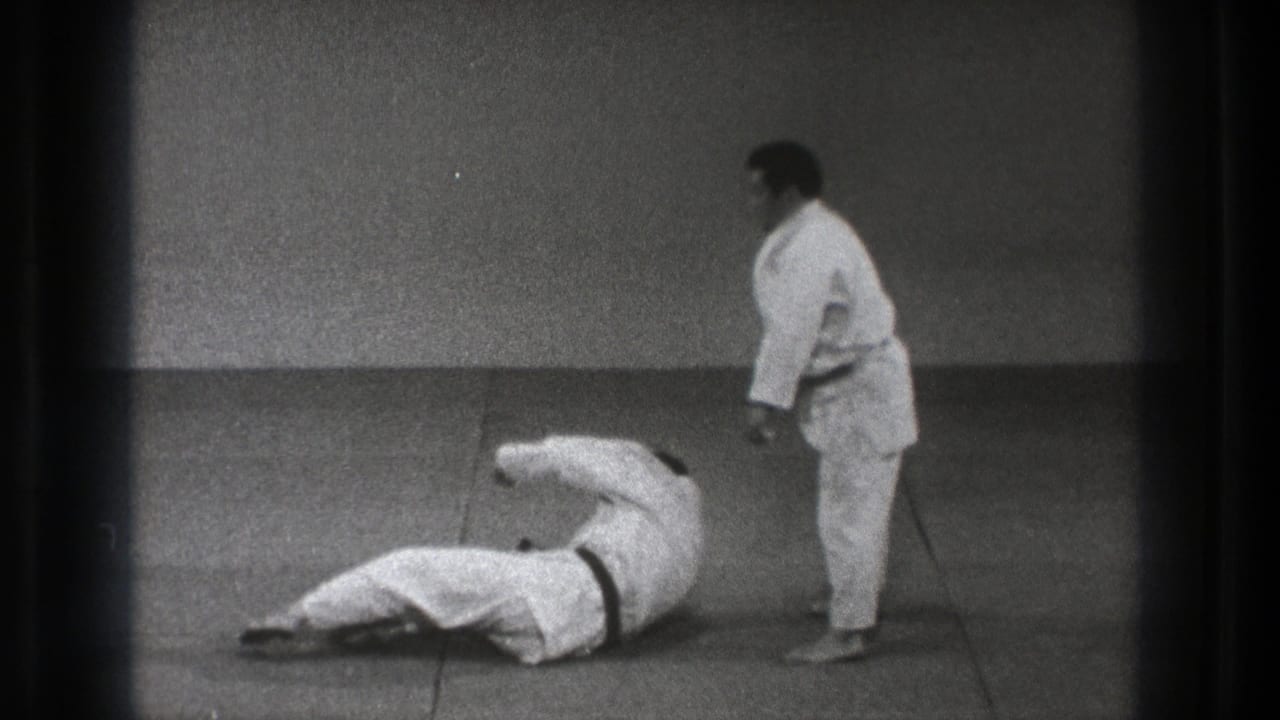 One martial arts practitioner lies on his side on the tatami as another stands nearby in the black-and-white Tokyo training footage.