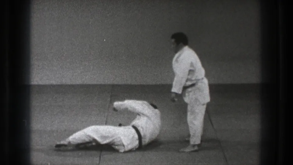 One martial arts practitioner lies on his side on the tatami as another stands nearby in the black-and-white Tokyo training footage.