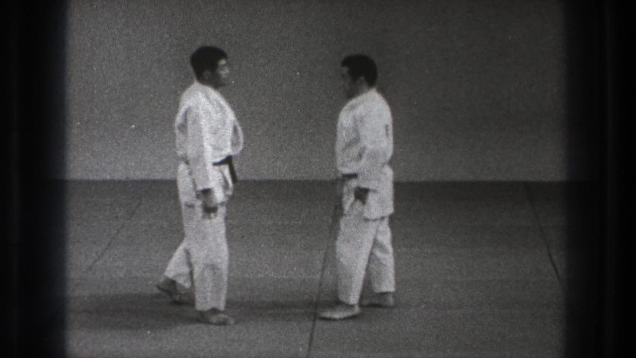 Two adult men in white karate gi face each other on a plain mat.