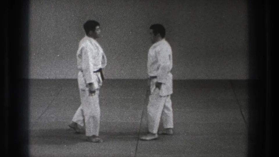 In 1971 Tokyo footage, two adult karate practitioners in white gi stand facing each other on a simple mat against a plain wall.