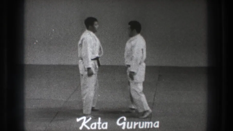 Two adult karate practitioners in white gi face each other as the handwritten title Kata Guruma appears over the 1971 Tokyo frame.