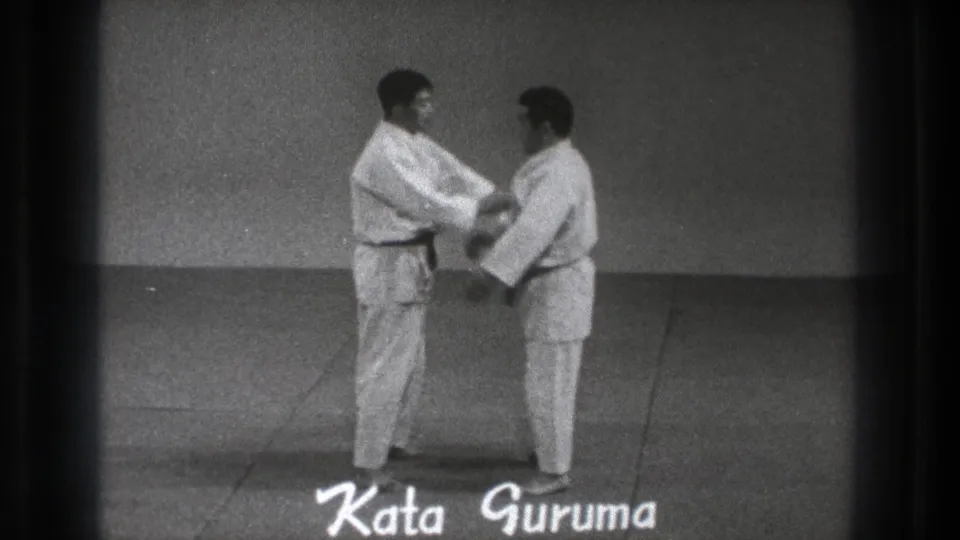 In the Tokyo karate demonstration, one adult man in a gi grips the other at close range during the Kata Guruma movement.