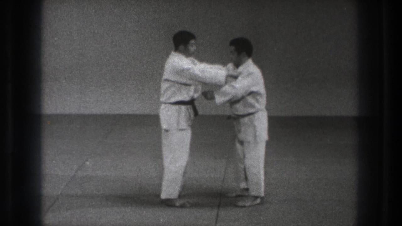Two adult karate practitioners continue the close-range Kata Guruma demonstration on a plain mat in black-and-white 8mm footage.