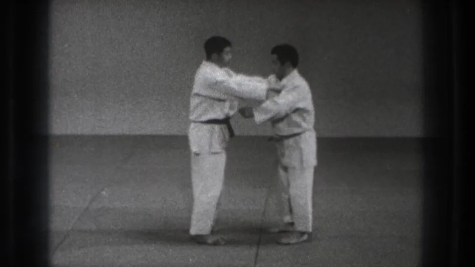 Two adult karate practitioners continue the close-range Kata Guruma demonstration on a plain mat in black-and-white 8mm footage.