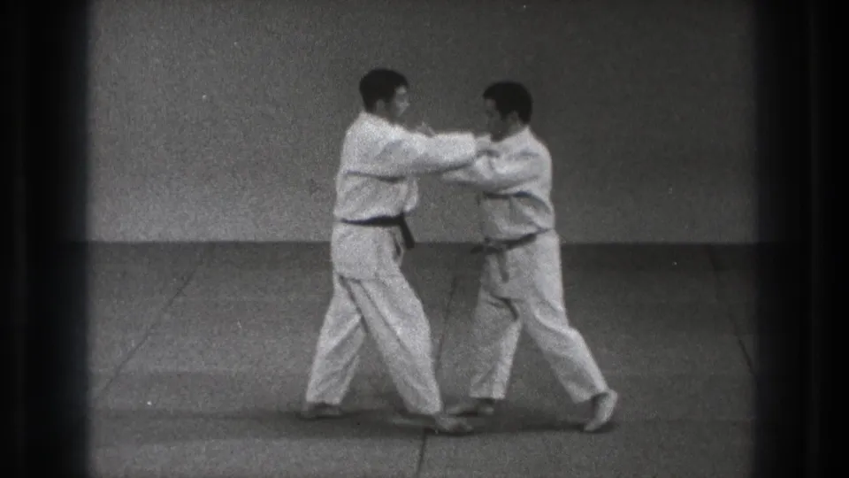 In the Tokyo 1971 karate practice footage, two adult men in white gi face each other on an indoor mat with their arms engaged.