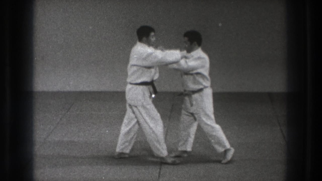 Two adult karate practitioners in white gi hold close arm contact while adjusting stance during the Tokyo practice sequence.