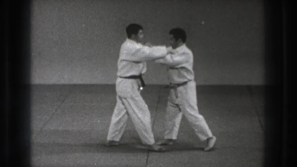Two adult karate practitioners in white gi hold close arm contact while adjusting stance during the Tokyo practice sequence.