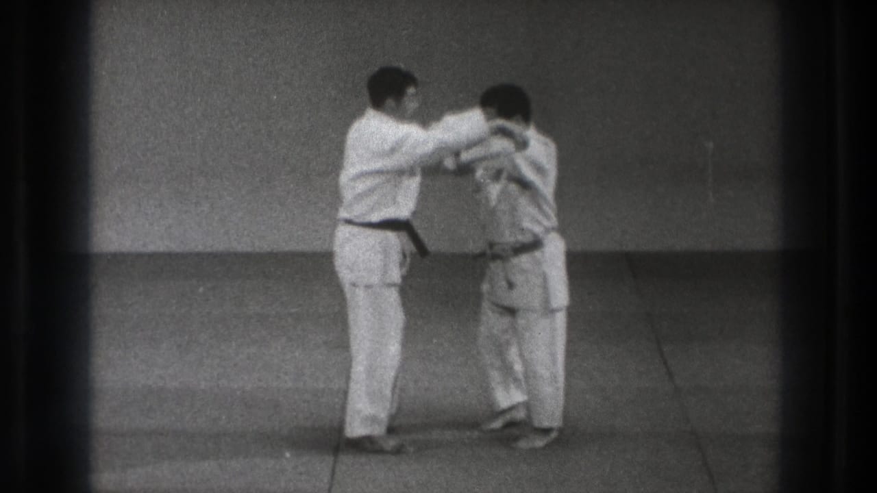 The black-belt practitioner appears to guide or control his partner's upper body during a karate technique in the indoor dojo setting.