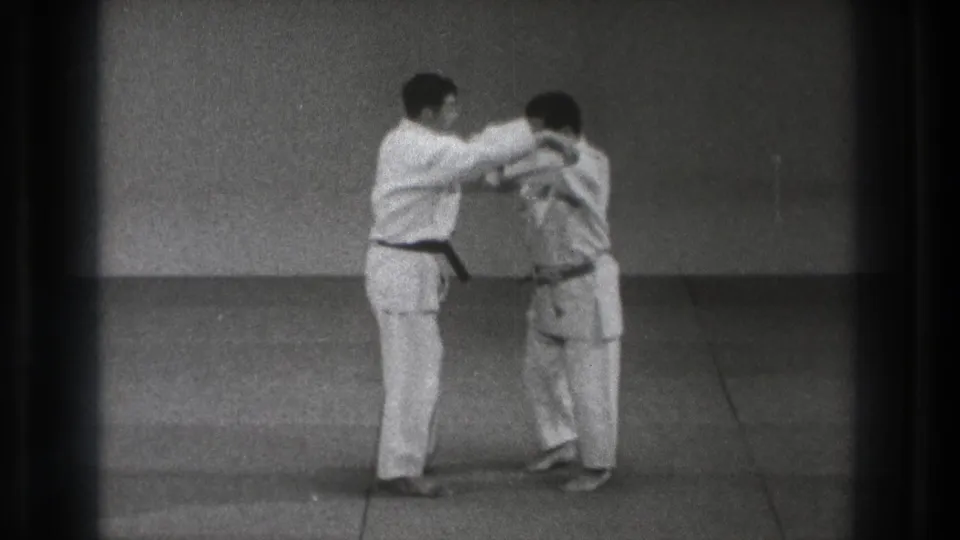 The black-belt practitioner appears to guide or control his partner's upper body during a karate technique in the indoor dojo setting.