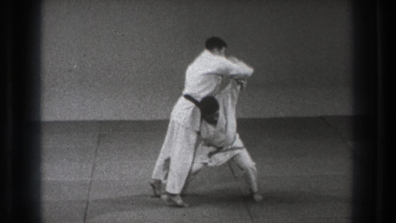 Two men in white martial arts uniforms begin a judo throw on a plain mat.