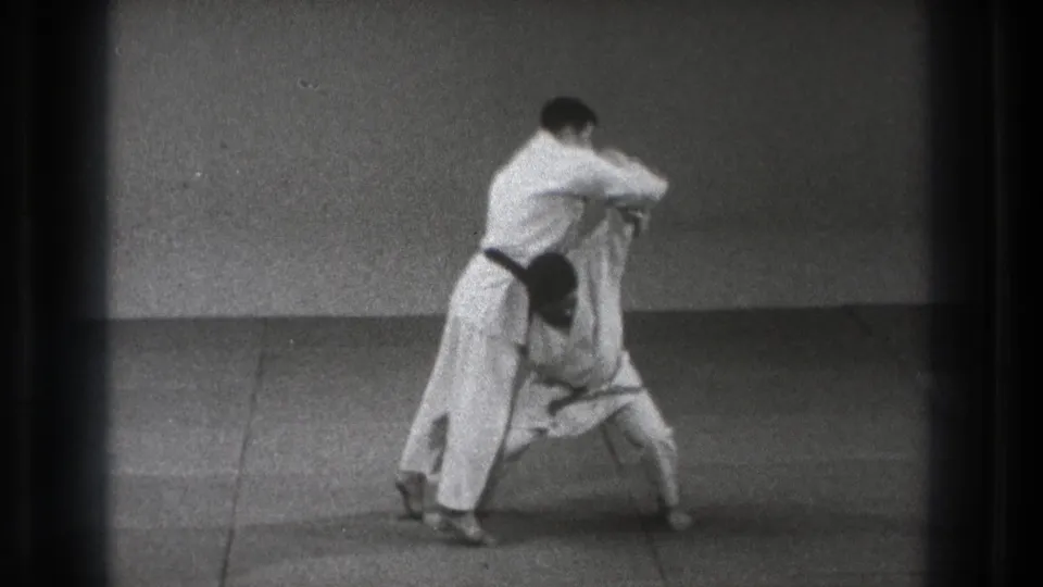 In Tokyo 1971 footage, two men in judogi engage at close range as one prepares to lift the other during a judo demonstration.