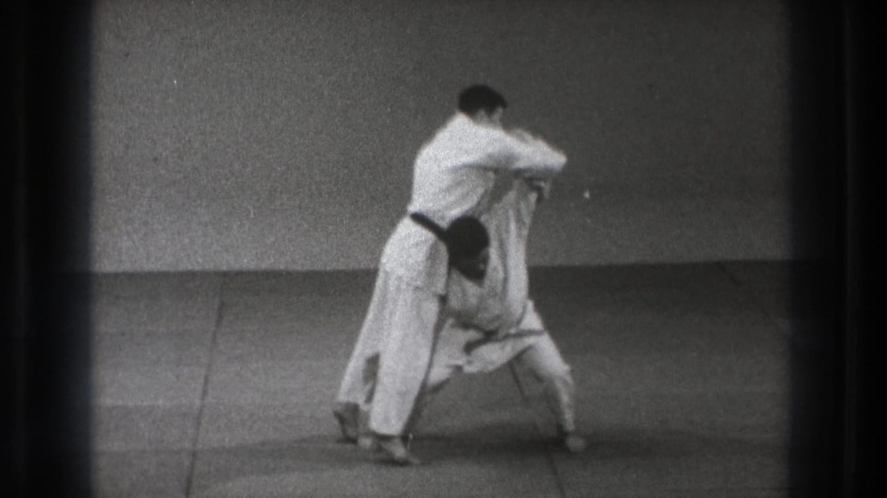 The 1971 Tokyo home-movie frame shows two men in white judogi locked together before the lift, filmed against a bare wall.