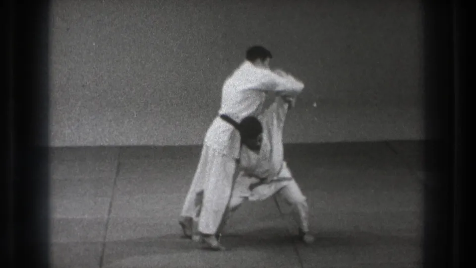 The 1971 Tokyo home-movie frame shows two men in white judogi locked together before the lift, filmed against a bare wall.