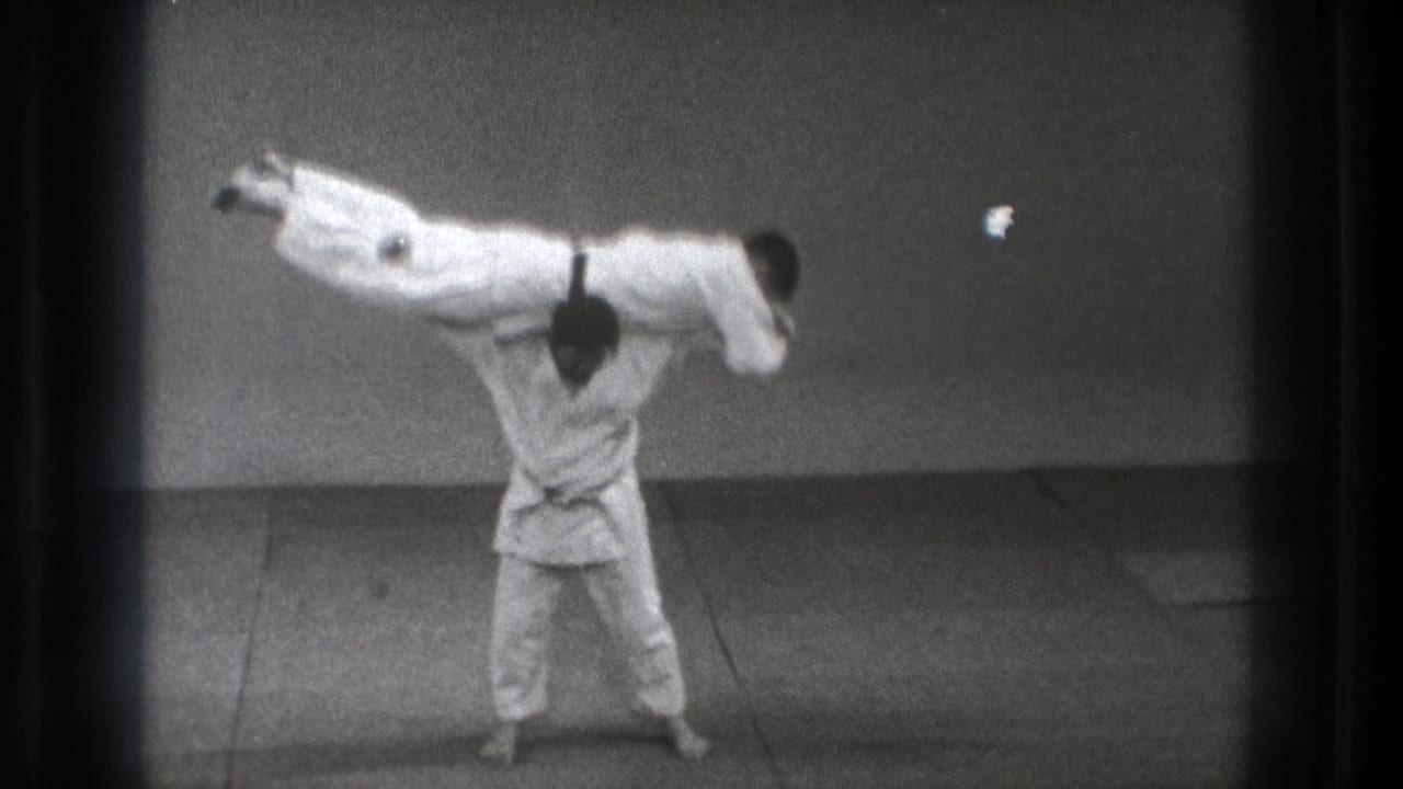 In 1971 Tokyo footage, a martial artist in a white gi carries another gi-clad person across his shoulders on an indoor mat.