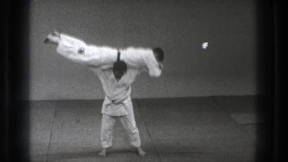 In 1971 Tokyo footage, a martial artist in a white gi carries another gi-clad person across his shoulders on an indoor mat.