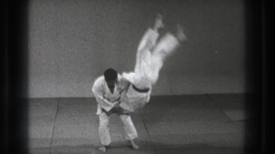 A martial artist in a white gi holds his partner as the partner rotates through the air during the throw on an indoor mat.