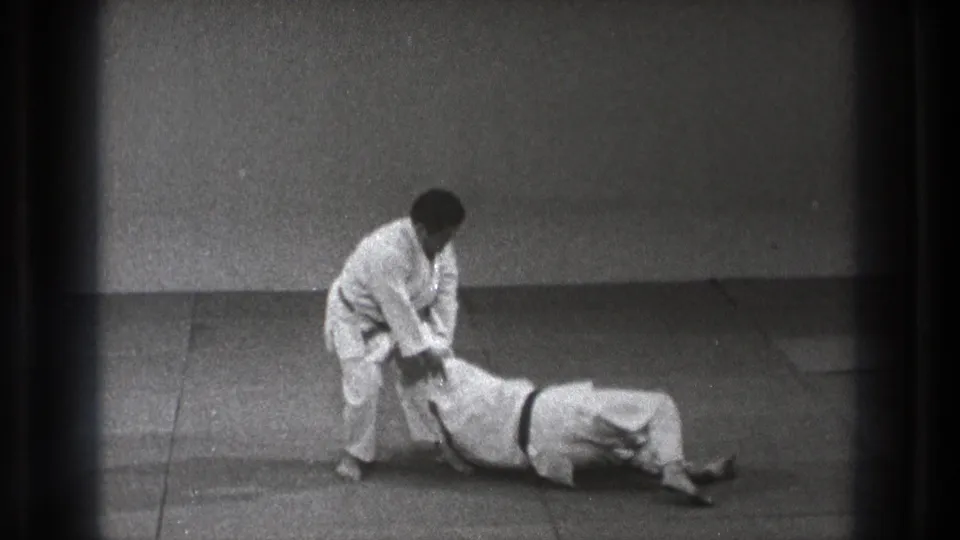 In Tokyo 1971 footage, two men in white judogi demonstrate a grappling position on an indoor mat, with one standing and one grounded.