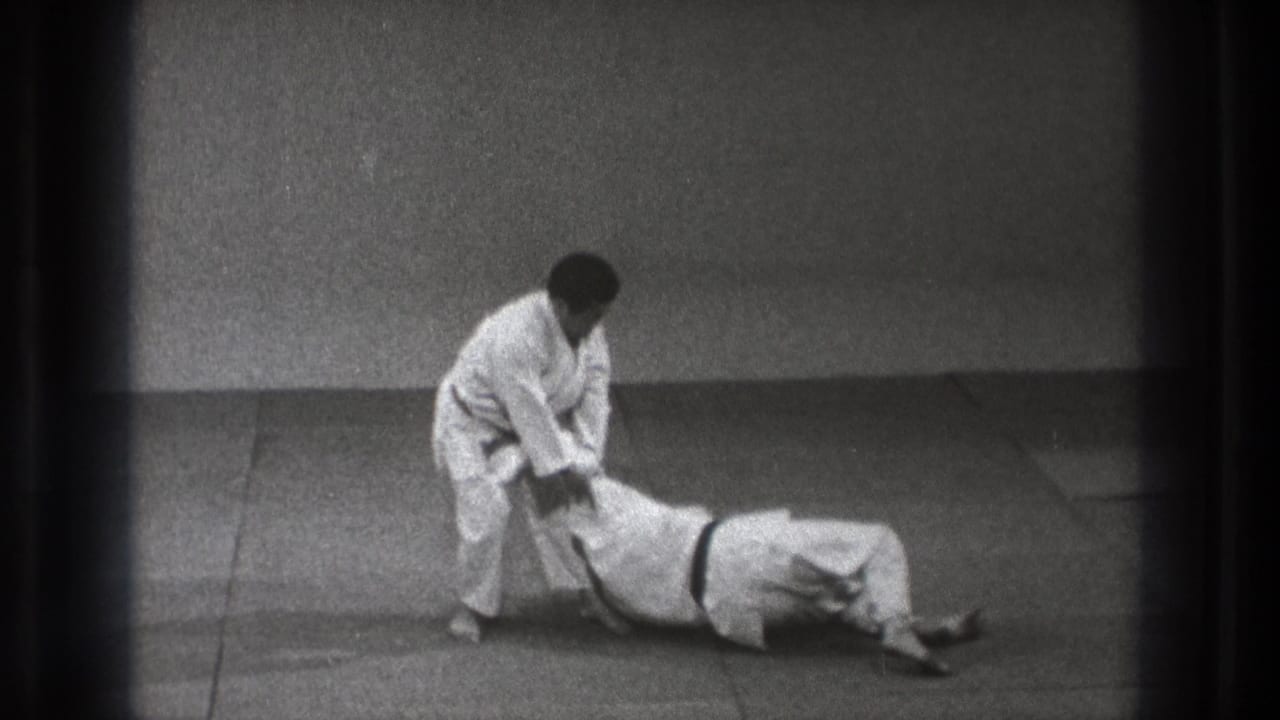 The 1971 Tokyo judo demonstration continues as the standing practitioner maintains an arm grip while the partner remains on the mat.