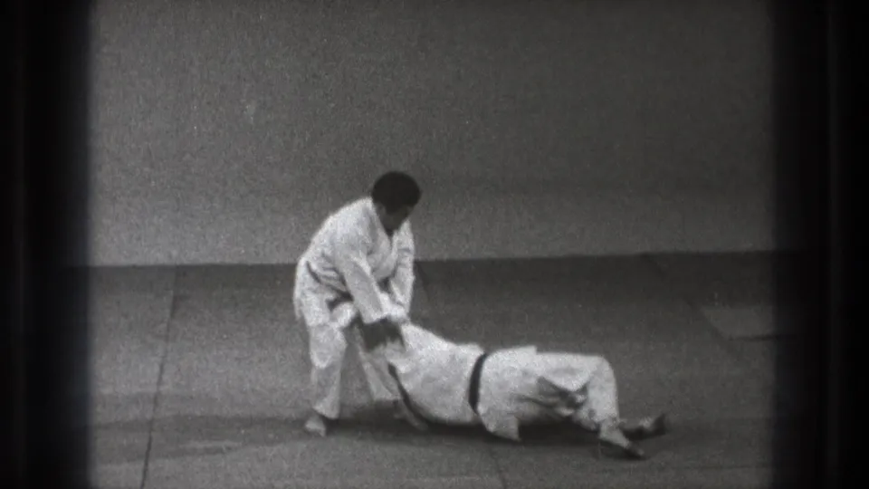 The 1971 Tokyo judo demonstration continues as the standing practitioner maintains an arm grip while the partner remains on the mat.