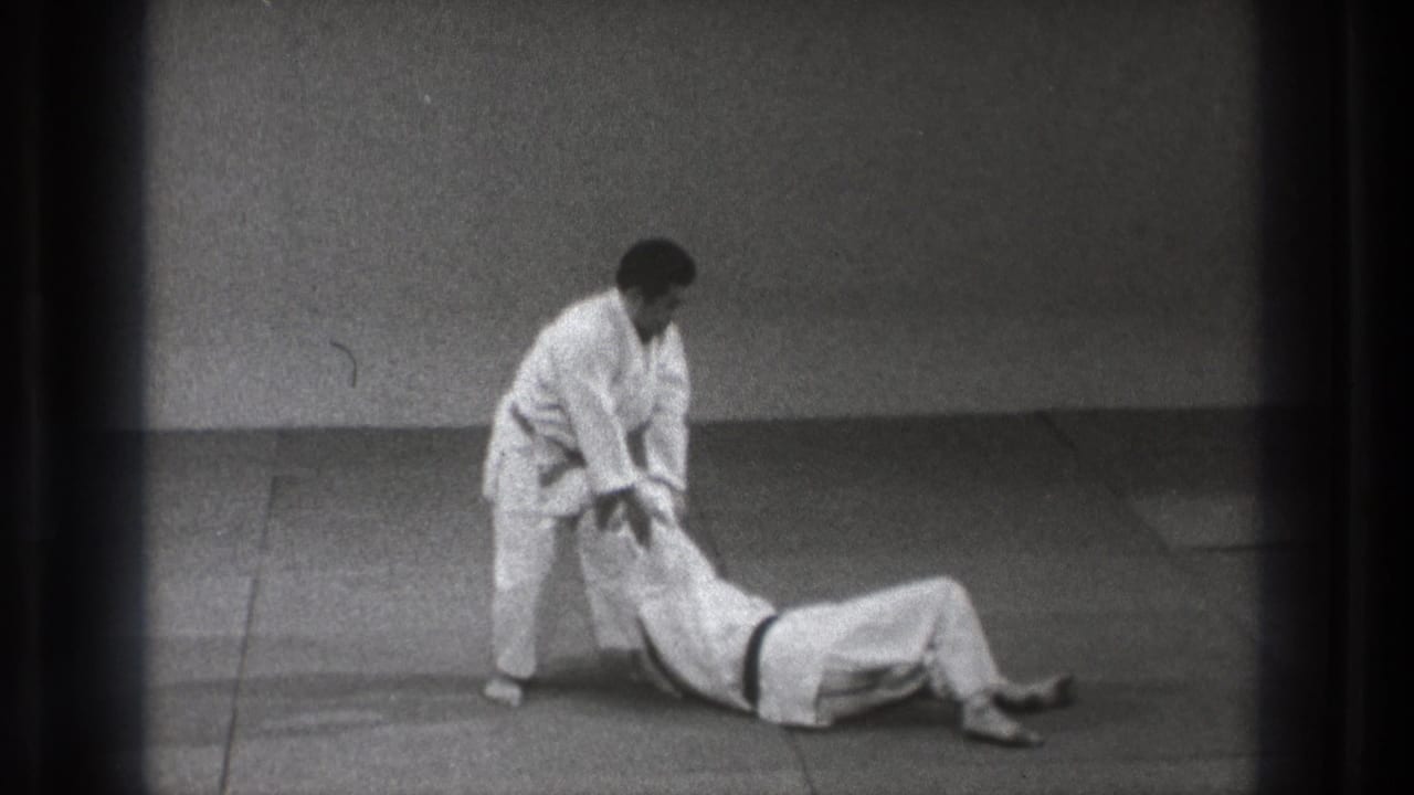 In the indoor judo sequence, the standing man keeps contact with his partner during the follow-through of a controlled grappling move.