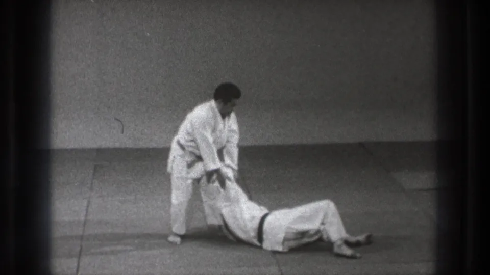 In the indoor judo sequence, the standing man keeps contact with his partner during the follow-through of a controlled grappling move.