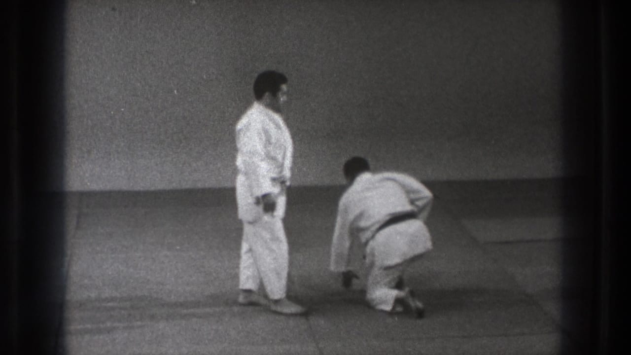 Two men in white gi on a dojo mat, one standing while the other kneels.
