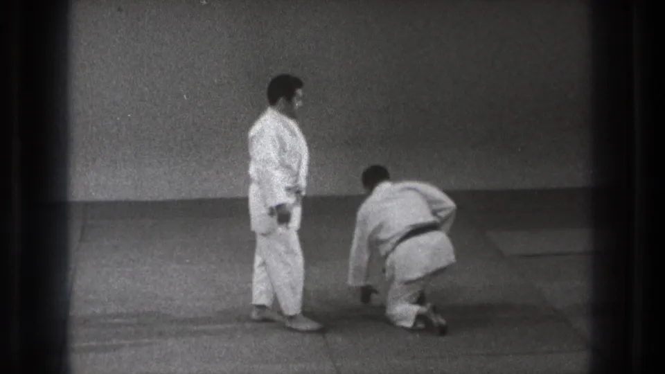 1971 Tokyo home-movie frame showing two martial arts practitioners on a mat, with one man kneeling and one standing nearby.