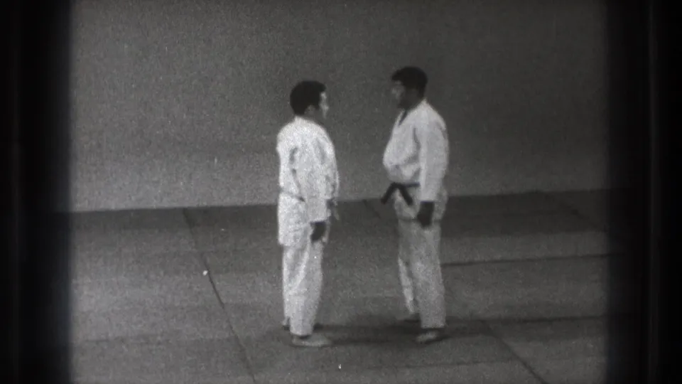 1971 Tokyo home-movie frame showing two men in gi bowing or leaning toward each other during dojo practice.