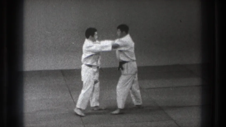 1971 Tokyo frame showing two martial arts practitioners making hand contact during a grip or pushing drill on the mat.